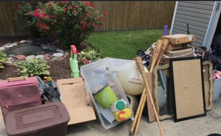 Assorted household items and yard supplies piled on a driveway, including plastic bins, cardboard boxes, wooden frames, and a pink cooler, with a green lawn and flowering shrubs in the background