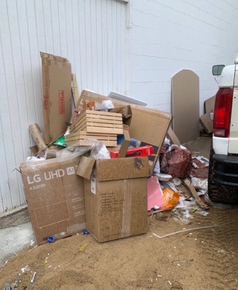 Pile of discarded cardboard boxes and debris scattered on dirt ground against a white corrugated metal building wall