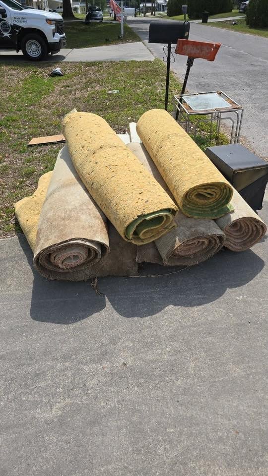 Rolled carpeting and rugs stacked on driveway during yard sale, with mailbox and residential street background