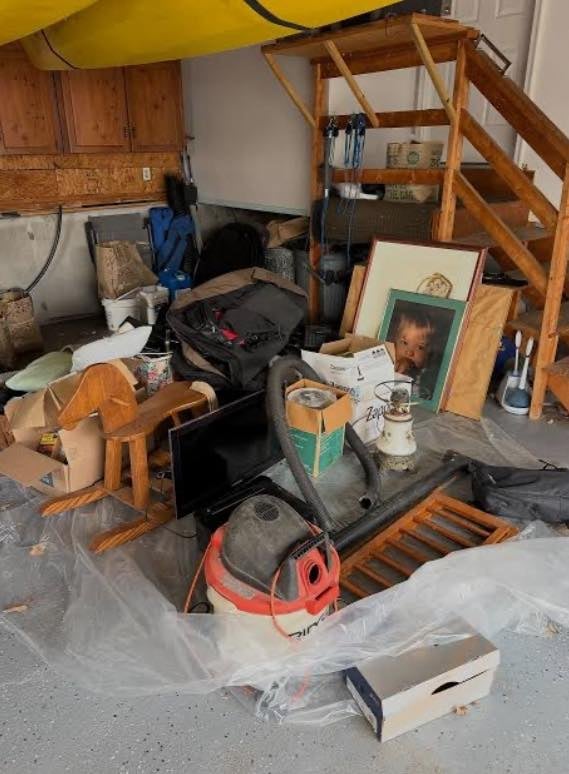 Crowded garage basement filled with miscellaneous items including wooden furniture, tools, boxes, and household clutter