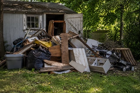 Dilapidated wooden shed surrounded by piles of discarded debris, scrap wood, and junk in an overgrown yard