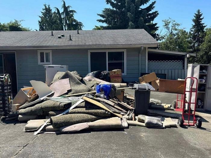 Pile of discarded wood, cardboard, and debris in a driveway in front of a small house with garage and evergreen trees